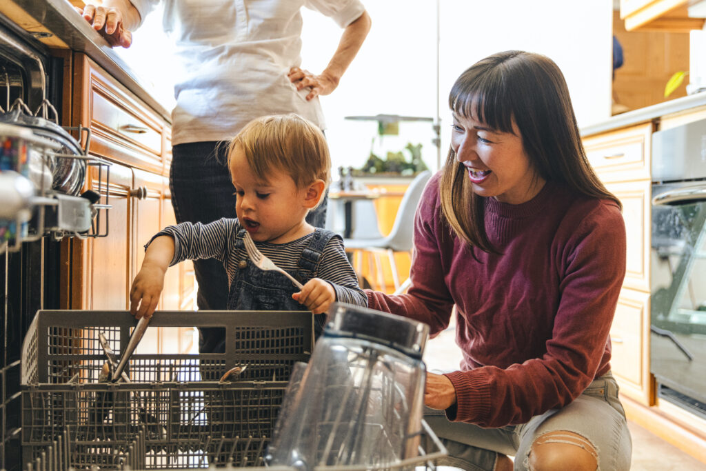 Par på kjøkkenet med småbarn. Kvinne sitter med barnet som holder å leke med ting i oppvaskemaskinen
